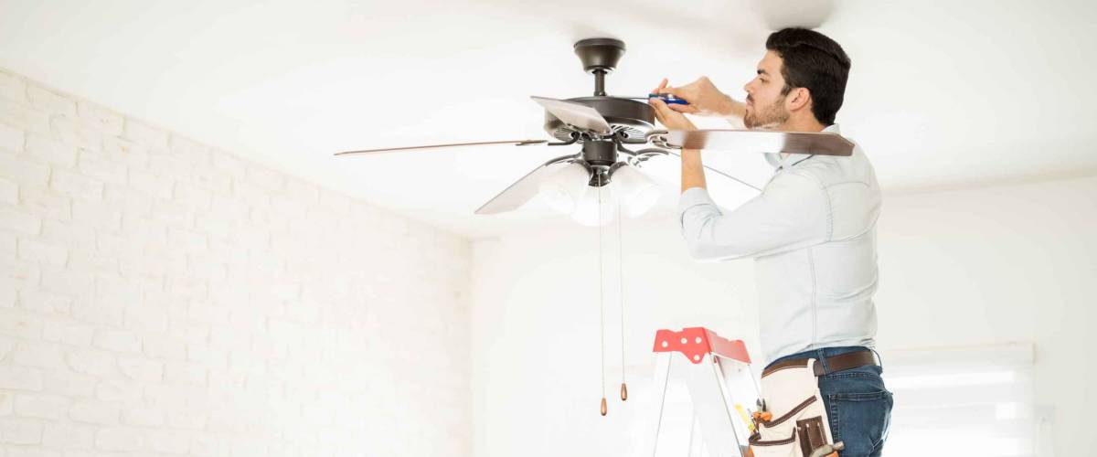 Profile view of a male electrician stepping on a ladder and installing a ceiling fan in a house