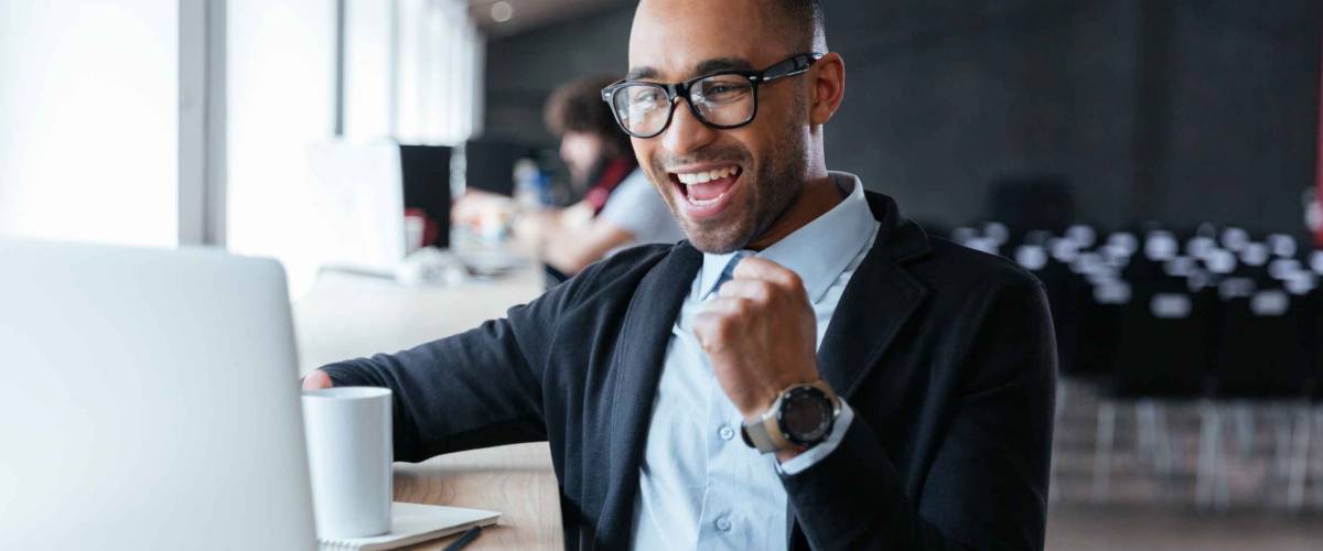 Handsome young businessman sitting at his ddesk, celebrating success with arms raised while looking at his laptop screen