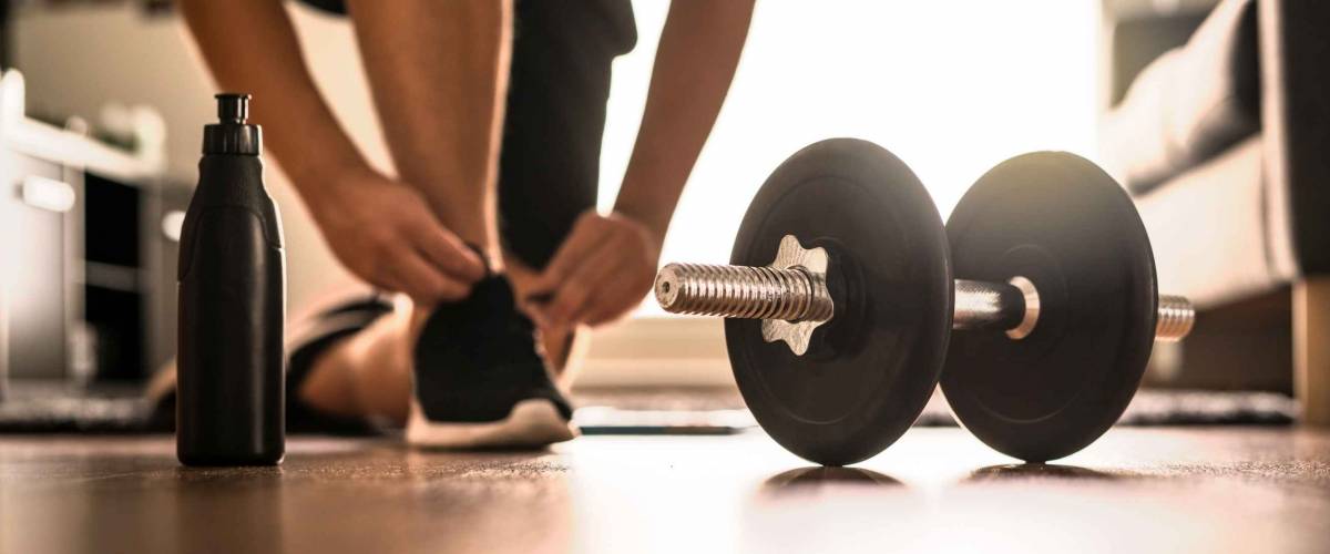 Morning workout routine in home gym. Fitness motivation and muscle training concept. Man in sneakers tying shoelaces in sunlight. Athlete starting exercise with dubbell weight.