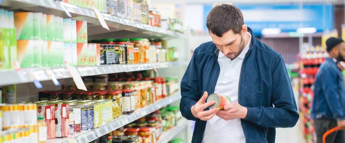 At the Supermarket: Handsome Man Uses Smartphone and Looks at Nutritional Value of the Canned Goods. He's Standing with Shopping Cart in Canned Goods Section.