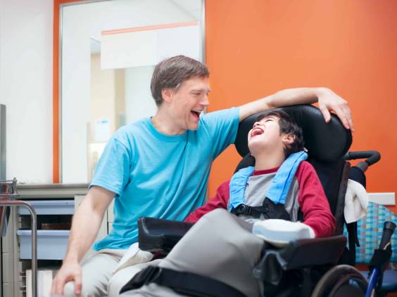 Father talking with disabled biracial son sitting in wheelchair while waiting in doctor's office, laughing together.