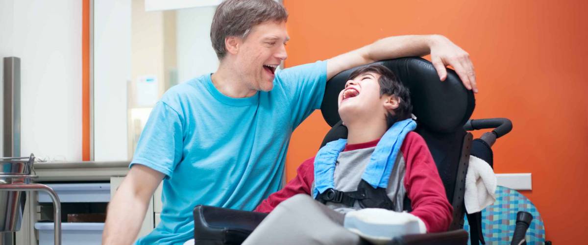 Father talking with disabled biracial son sitting in wheelchair while waiting in doctor's office, laughing together.