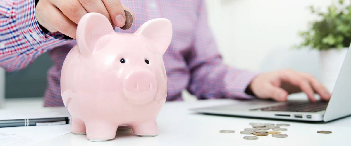 Man putting coin into a piggy bank, while sitting at his computer