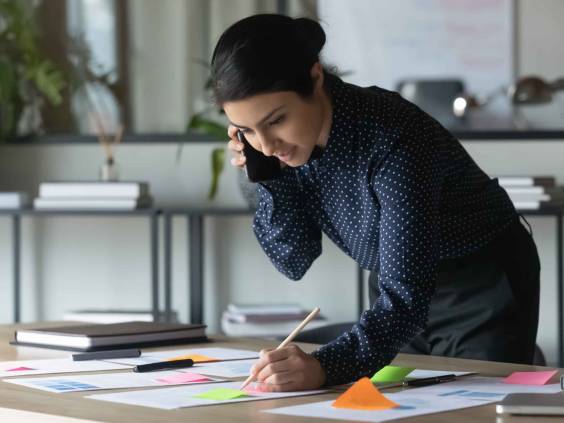 Serious Indian businesswoman talking on cellphone, writing notes on colorful sticky papers in office.