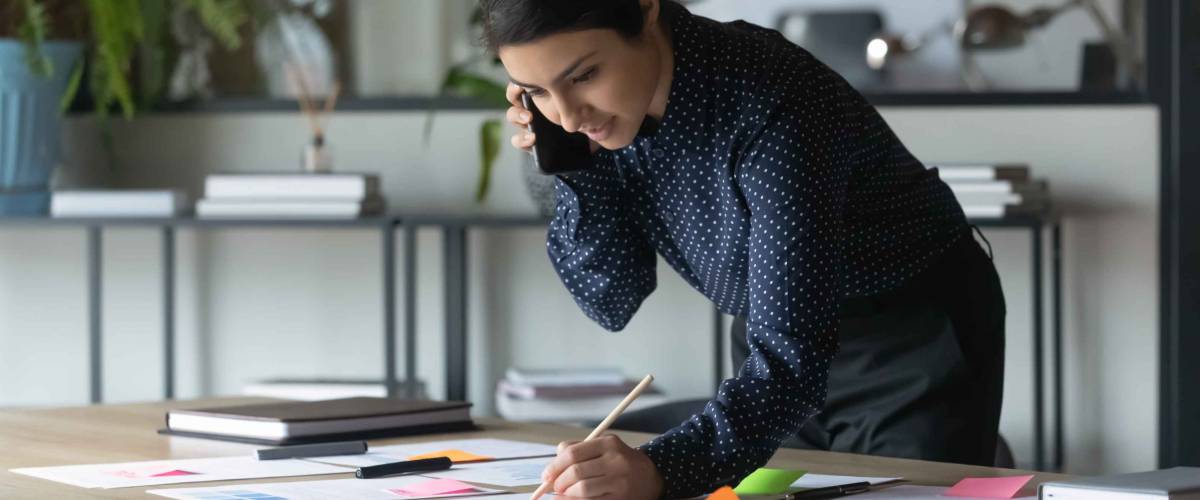 Serious Indian businesswoman talking on cellphone, writing notes on colorful sticky papers in office.