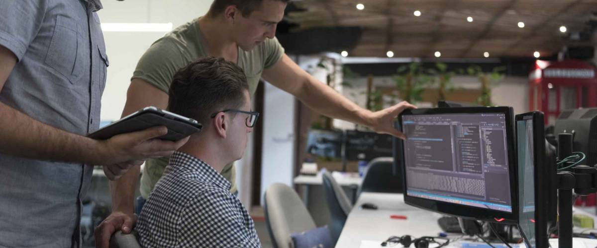 Three men standing in front of computer screen looking at code