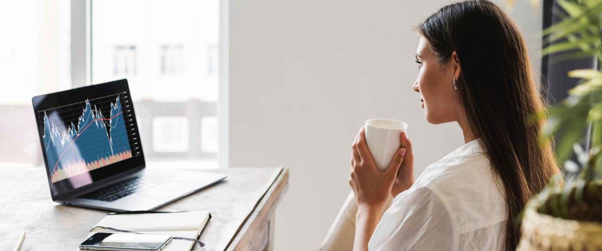 Attractive business woman sits at table in front of laptop and uses smartphone for work. Beautiful brunette girl analyzes stock market while at home. Millennial women on Internet.