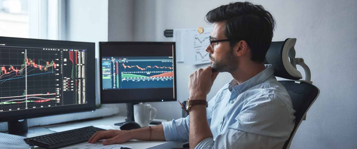 Busy working day. Side view of successful trader or businessman in formal wear and eyeglasses working with charts and market reports on computer screens in his modern office