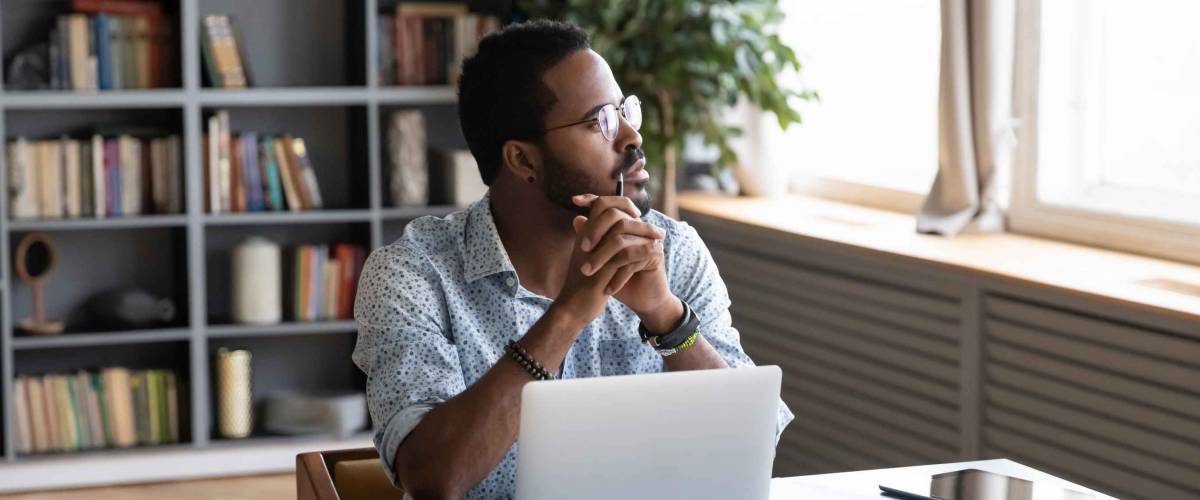 Thoughtful businessman sitting at table with computer, looking away.