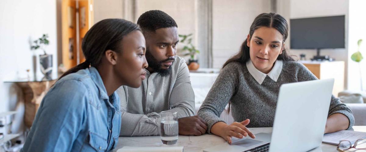 Financial planner going over savings plans on a laptop with a young couple at a table in their living room at home