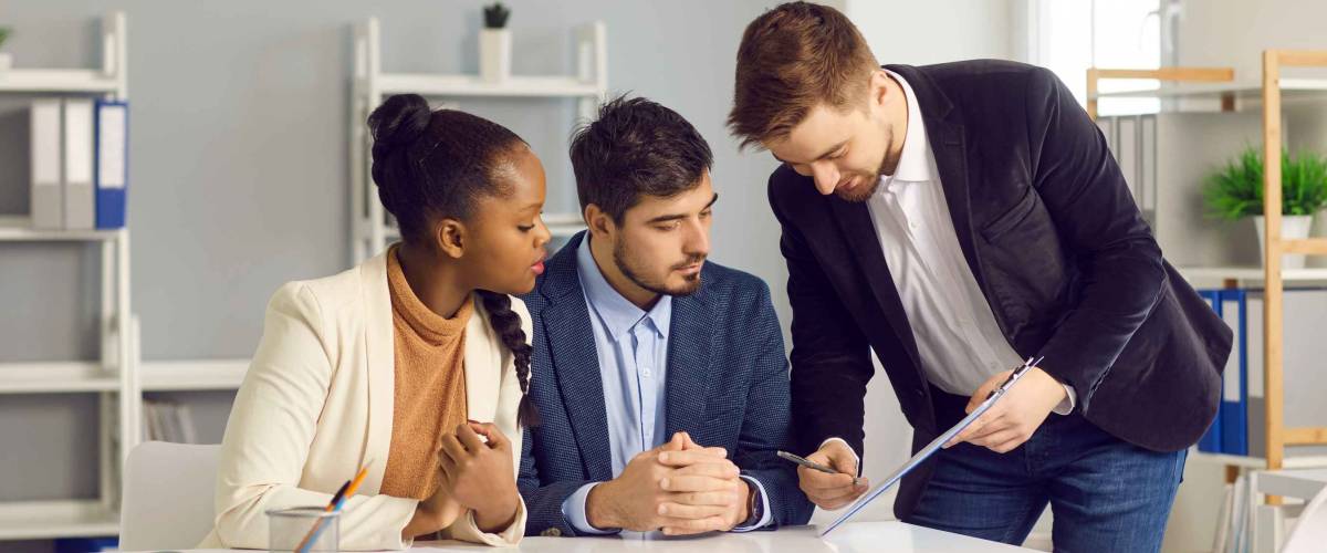 Bank employee gives documents for signature to a Caucasian man and his dark-skinned wife. Specialist in lending and financial assistance for young families indicates the place of signature.