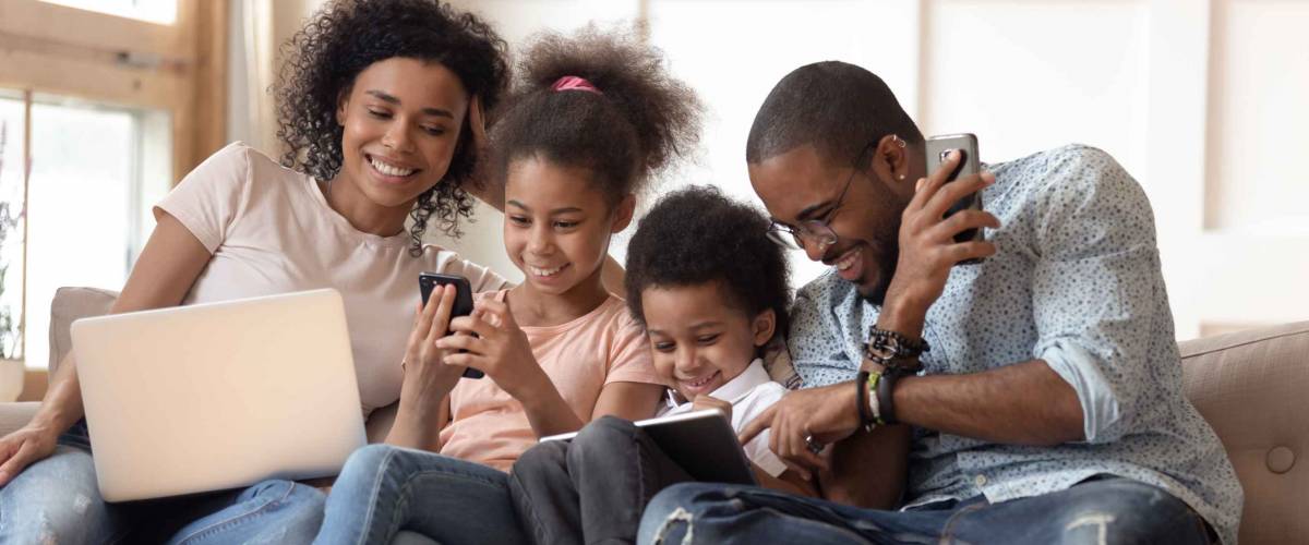 Family sits together on couch looking at devices, smiling.