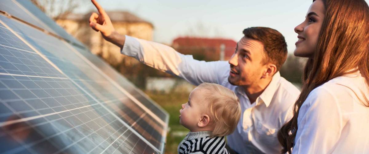 Man shows his family the solar panels on the plot near the house during a warm day. Young woman with a kid and a man in the sun rays look at the solar panels.