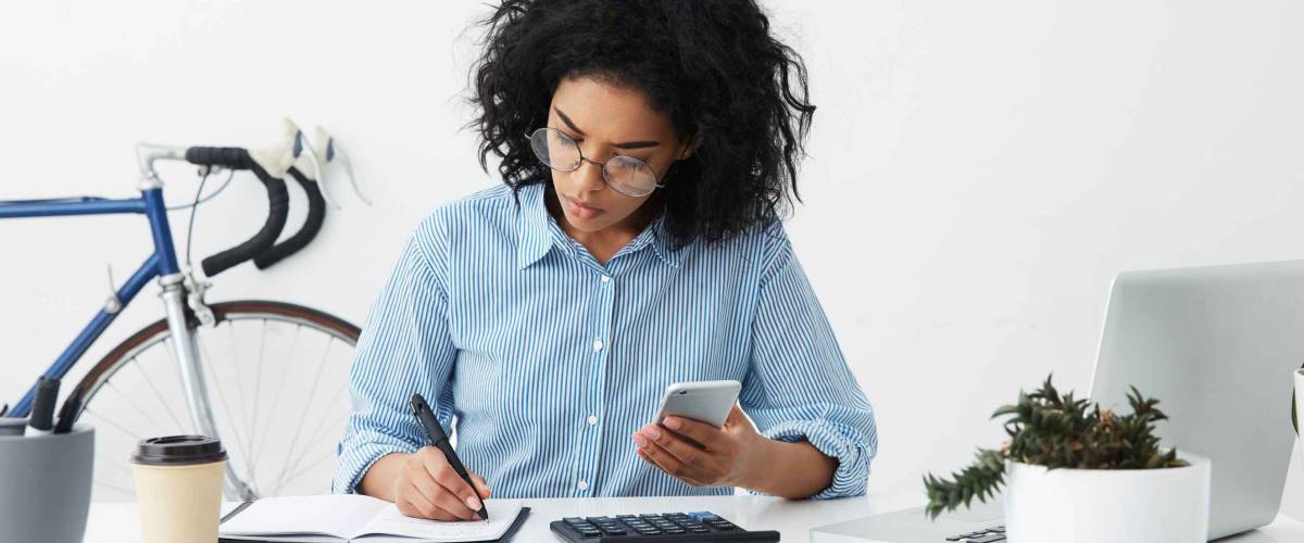Concentrated focused Afro American woman holding phone in one hand and making notes with pen in other