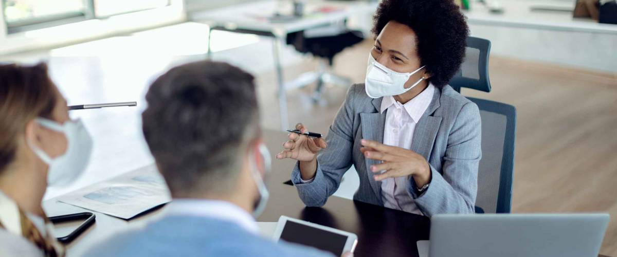Happy black financial advisor communicating with a couple on a meeting in the office. All of them are wearing protective face masks due to COVID-19 pandemic.