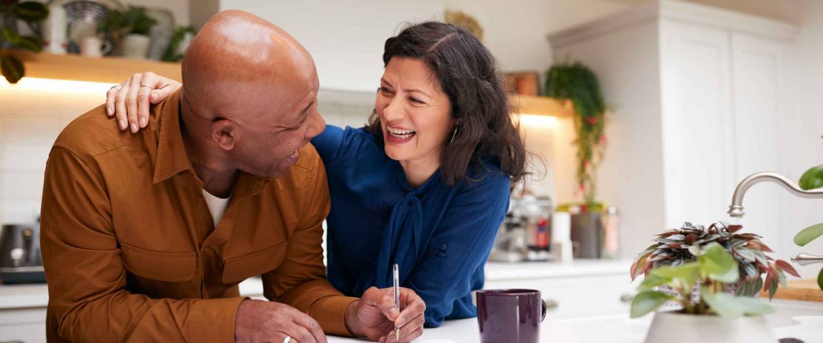 Mature Couple Reviewing And Signing Domestic Finances And Investment Paperwork In Kitchen At Home