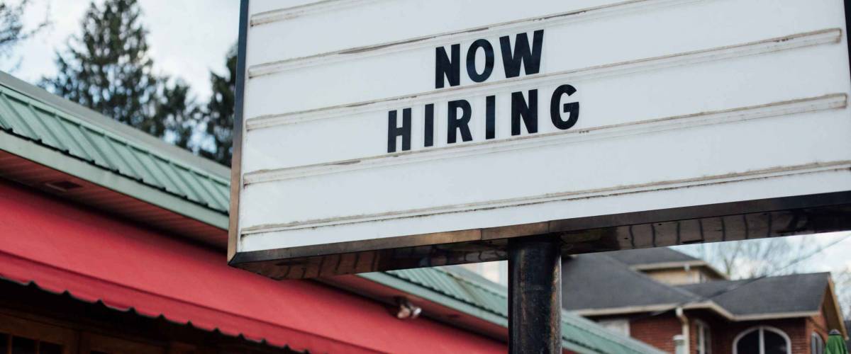 Typical american now hiring sign in black letters over the white background with local small business building