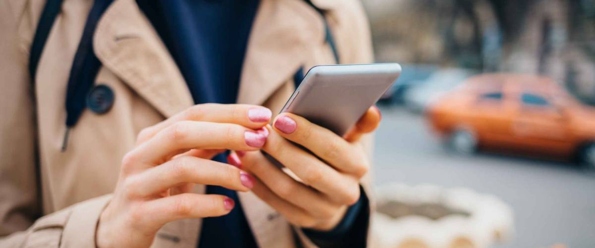 Young woman wearing beige coat using smart phone standing near road with cars in city at spring day. Close-up female's hands holding mobile phone outdoors.