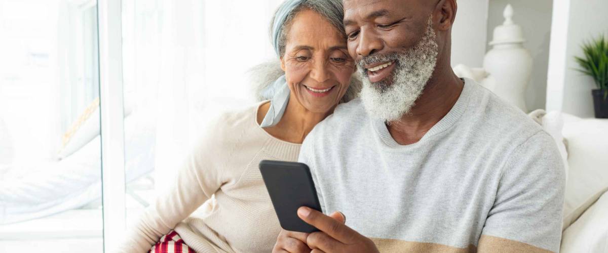 Front view of happy diverse senior couple using a smartphone on sofa in beach house. Authentic Senior Retired Life Concept