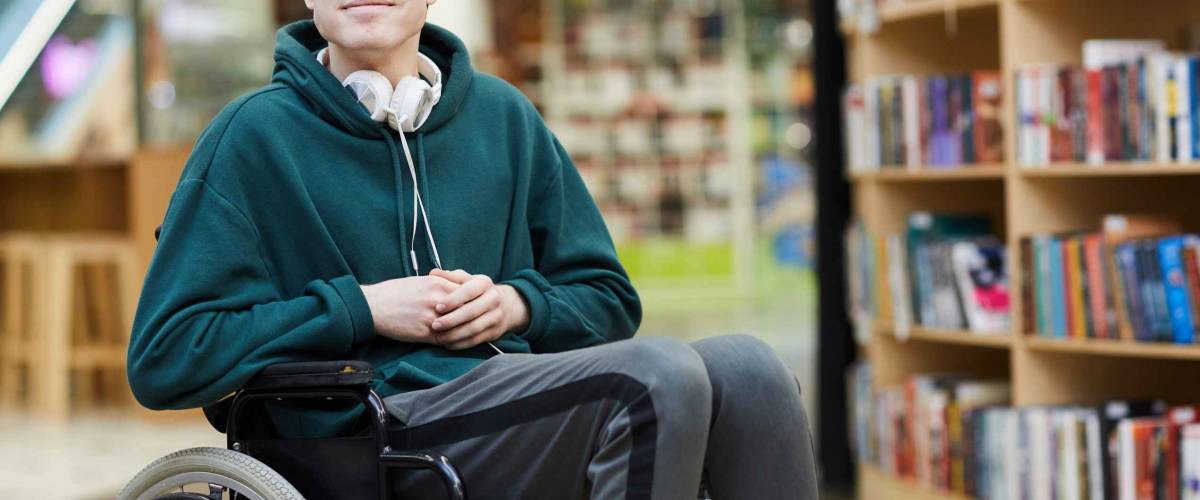 Content handsome young disabled student with headphones on neck siting in wheelchair and looking at camera in modern library or bookstore