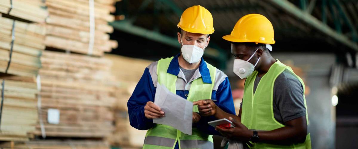 Two manual workers with face masks going though checklist while working in timber warehouse.