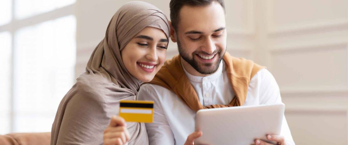 Online Shopping. Happy Muslim Couple Using Digital Tablet And Credit Card Buying And Purchasing Things Sitting On Sofa At Home. Joyful Customers, Modern Shopping And Sales Concept