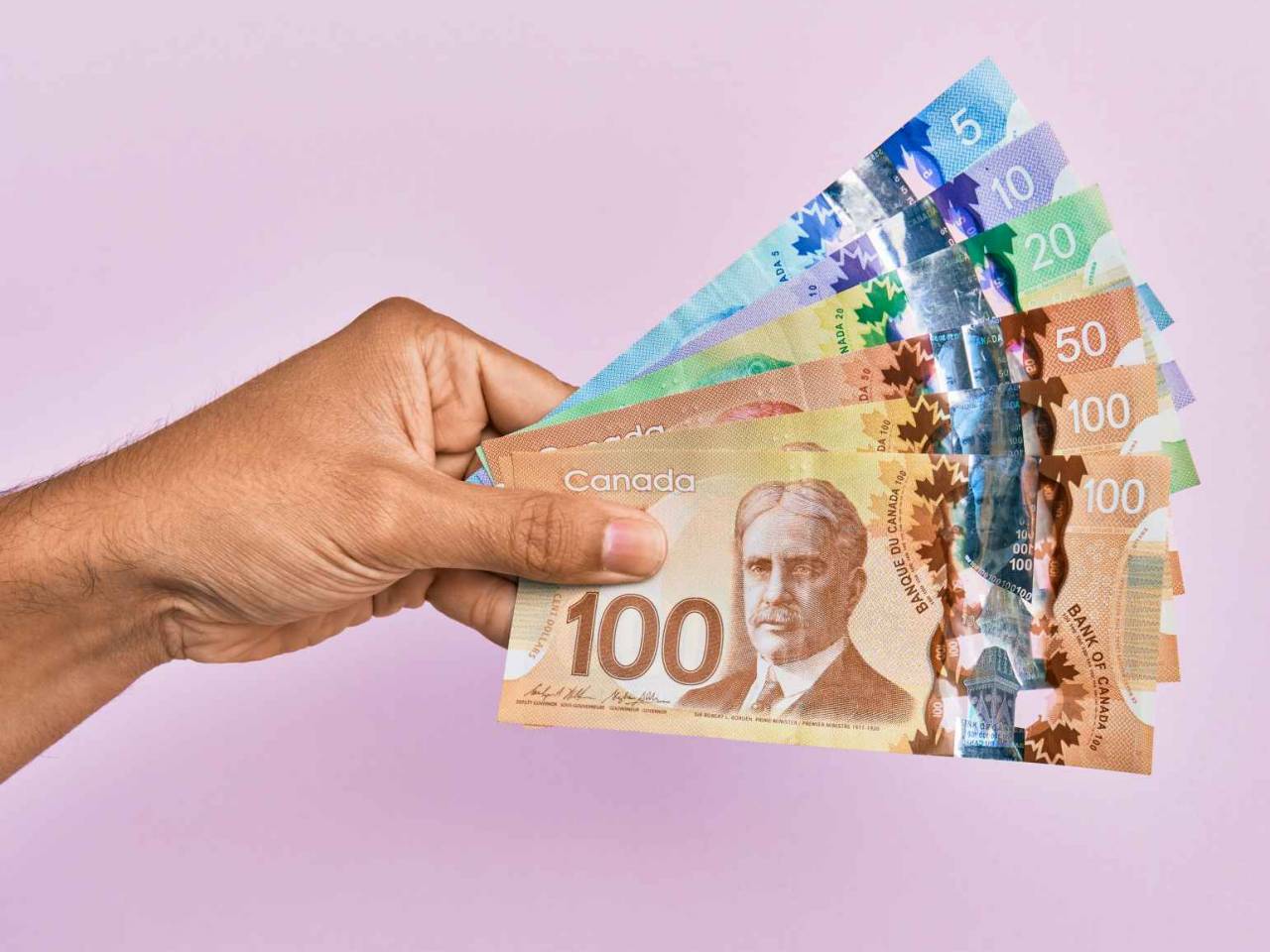 Hispanic hand holding canadian dollars banknotes over isolated pink background.