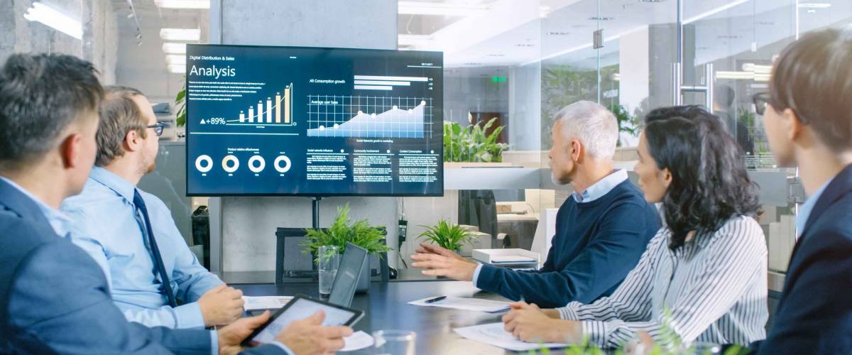 Group of people look at screen while sitting at boardroom table.