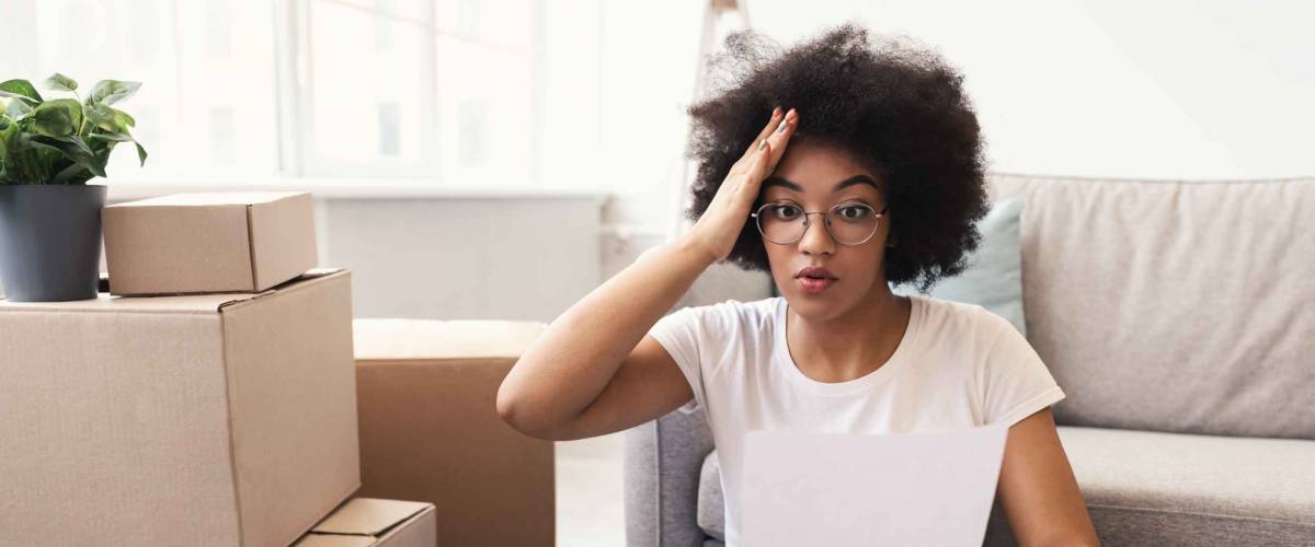 House Ownership And Rent Problem. Shocked African Woman Reading Paper Bill Or Sale Agreement Sitting Among Moving Boxes Indoors.
