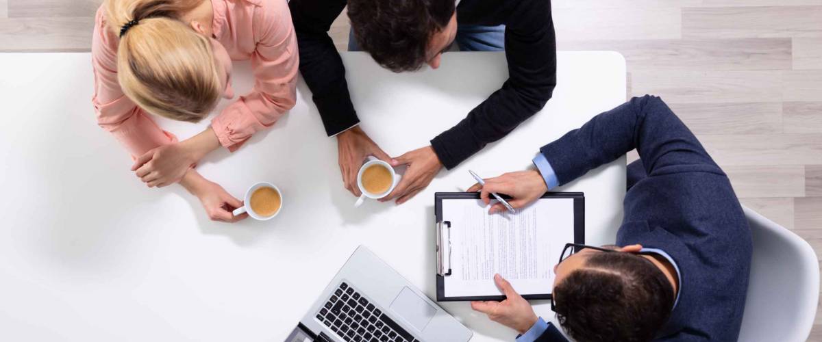 Happy Young Couple Consulting Male Friendly Financial Advisor With Coffee Cups At Office Desk