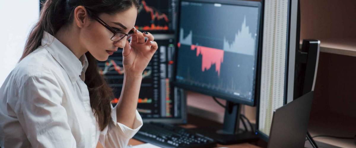Touching the glasses. Woman working online in the office with multiple computer screens in index charts.
