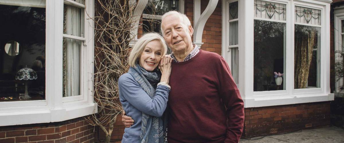 Senior couple are smiling for the camera while standing in front of their home.