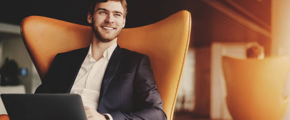 Young smiling successful man entrepreneur in formal business suite with a beard sitting on orange armchair with laptop in luxury office interior