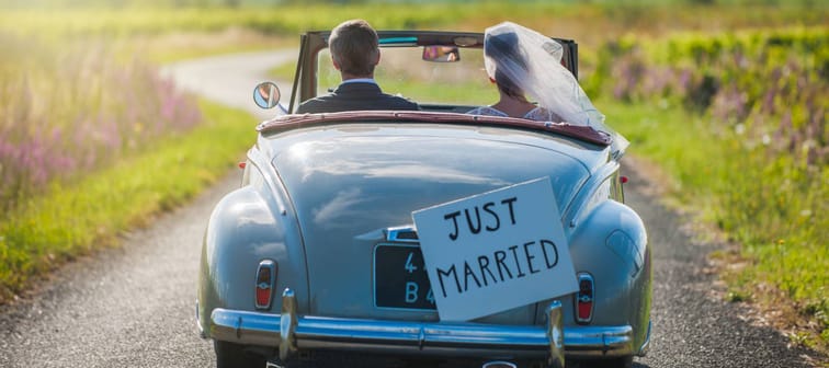A newlywed couple is driving a convertible retro car on a country road for their honeymoon, rear view