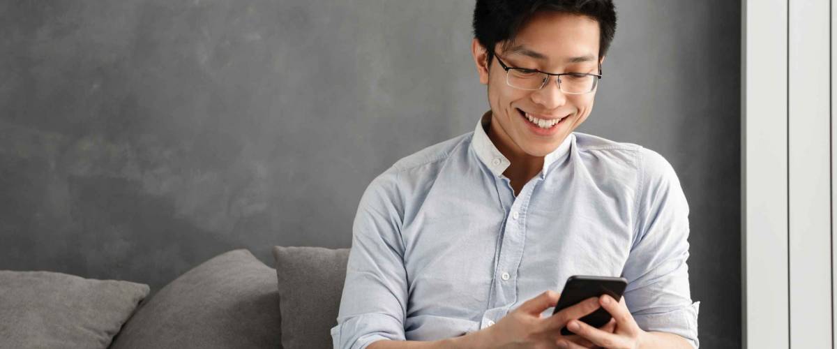 Portrait of a happy young asian man using mobile phone while sitting on a couch at home