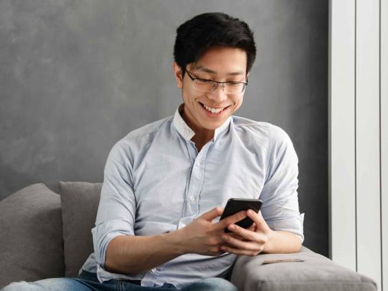Portrait of a happy young asian man using mobile phone while sitting on a couch at home