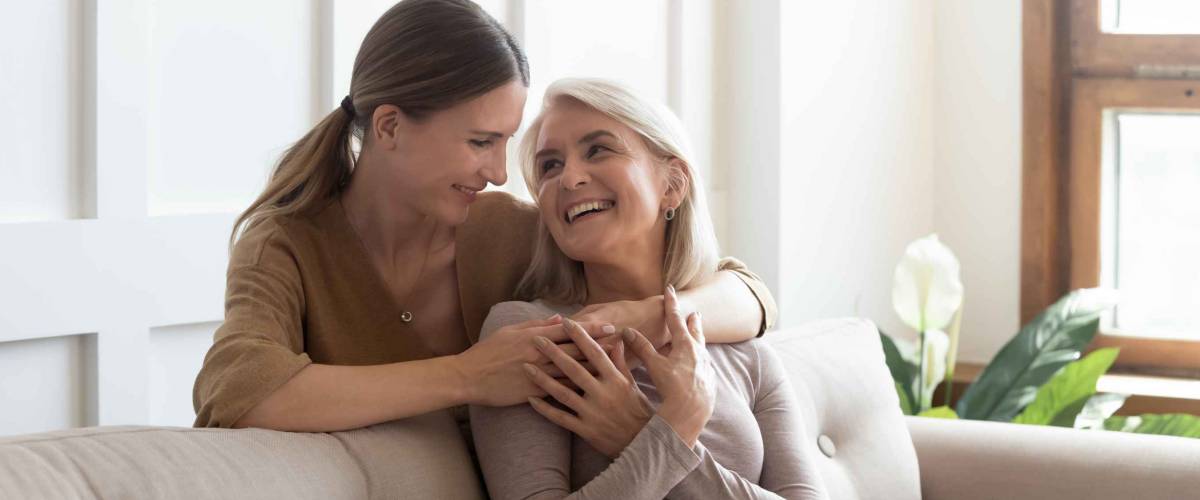 Loving adult daughter hugging older mother, standing behind couch at home