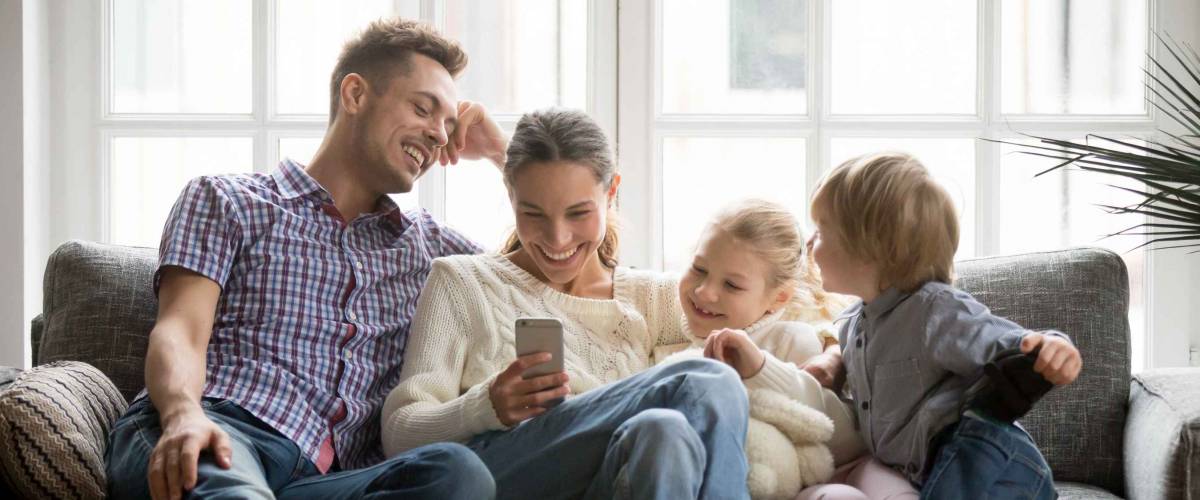 Cheerful young family with kids laughing looking at smartphone on couch