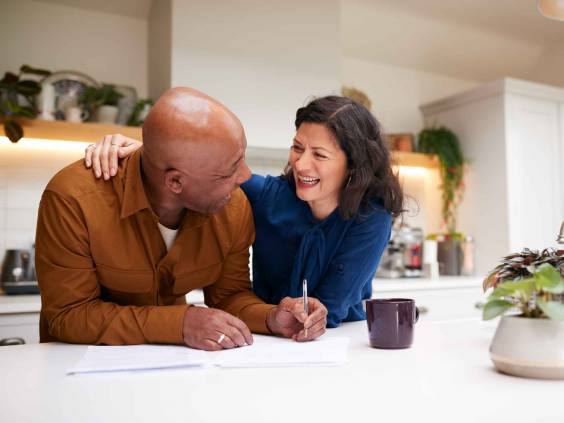 Mature Couple Reviewing And Signing Domestic Finances And Investment Paperwork In Kitchen At Home