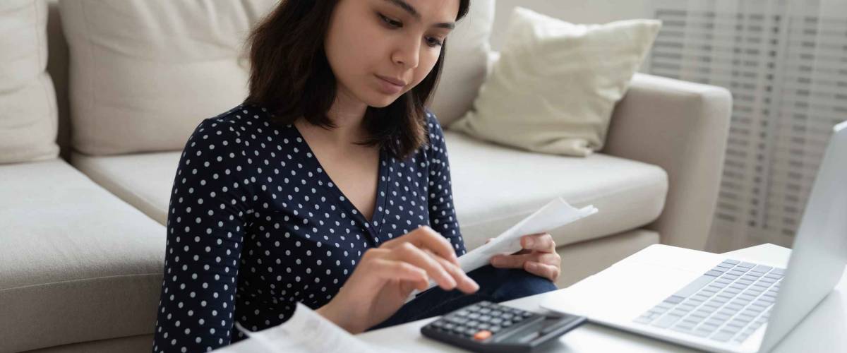Concentrated millennial vietnamese female engaged in home banking using calculator and laptop.