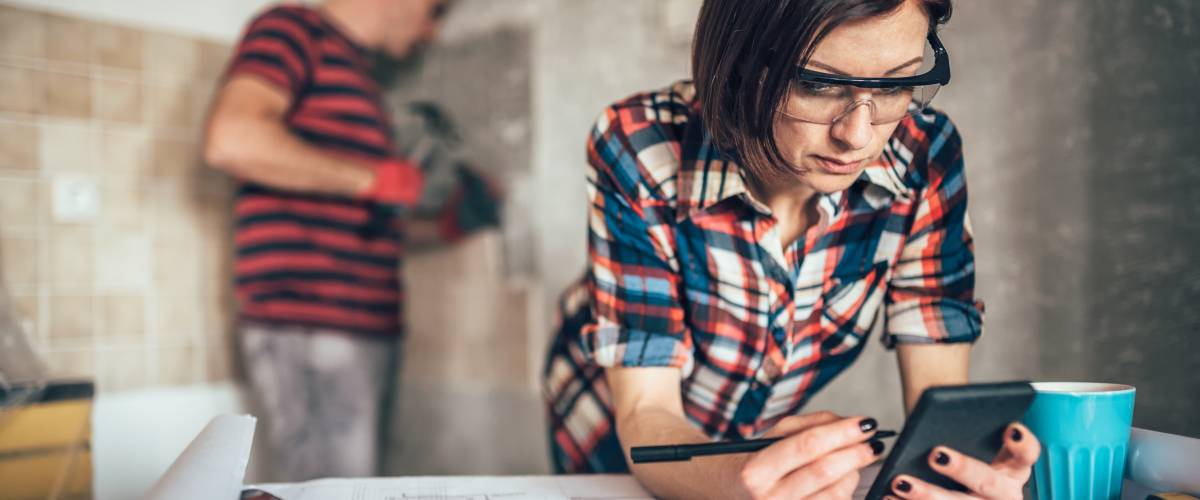 Woman using smart phone on the table with home reno tools at hand while her someone else removes old wall tiles in the kitchen