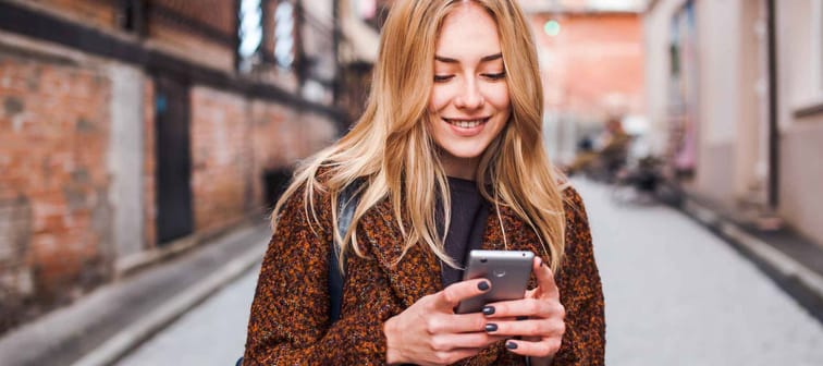 Young beautiful blonde and dreamy woman wearing casual terracotta coat writes sms online with smartphone chatting at street in autumn