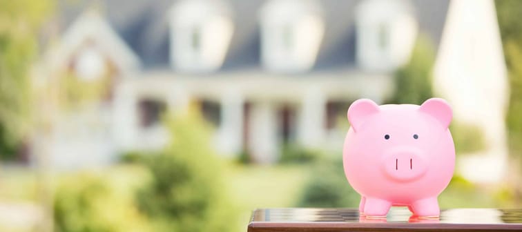 Piggy bank in front of a house, house is blurred, implying focus on savings over mortgage payoff.
