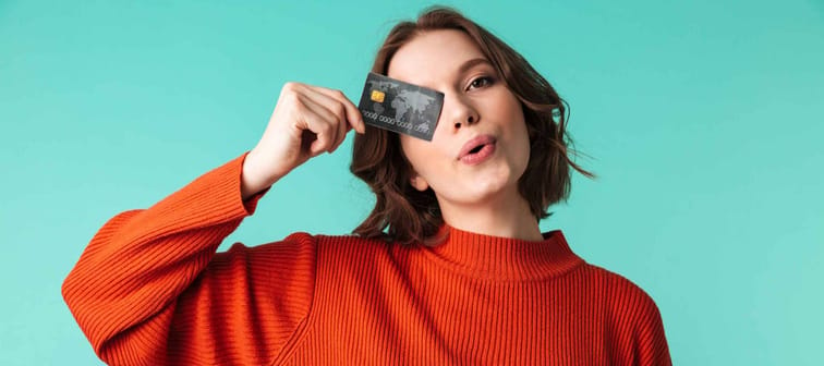 Portrait of a pretty young woman dressed in sweater holding credit card at her face isolated over blue background