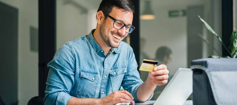Happy cheerful smiling young adult man doing online shopping or e-shopping satisfied entrepreneur making online payment paying for service or goods collecting fee paying happy