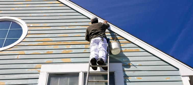 A contractor or painter on a ladder doing exterior paint work, sanding, trim work and repairs on a house, condo or building with wood siding.
