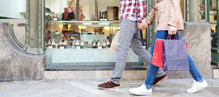 Lower body section of a young tourist couple walking by store windows and holding paper shopping bags in a destination city.