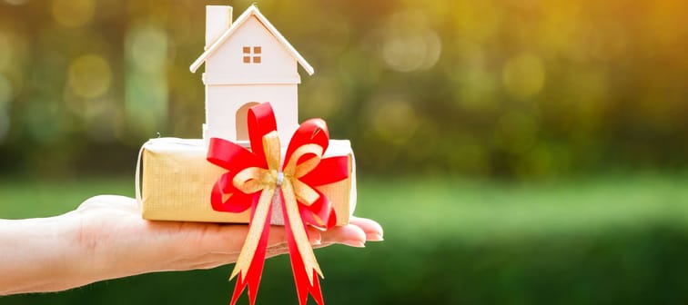 Woman hand holding a home model and gift box tied with red ribbon in the public park, for buying a new house or a real estate for family or person one loved concept.