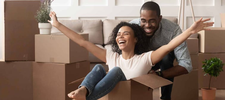 Young  Black couple first time home buyers having fun unpacking laughing on moving day, excited wife riding sitting in cardboard box while black husband push it in new house apartment